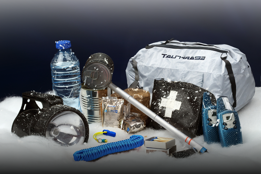 A collection of emergency kits sits upon fake snow includes: paracord, snow probe, tent bag, first aid kit, flashlight, emergency rations, walkie talkies, water, a whistle, and other supplies.