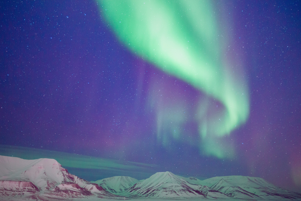 Over a snowy mountain range, a night sky stretches showing stars and aurora borealis of green, blue, purple, and white.