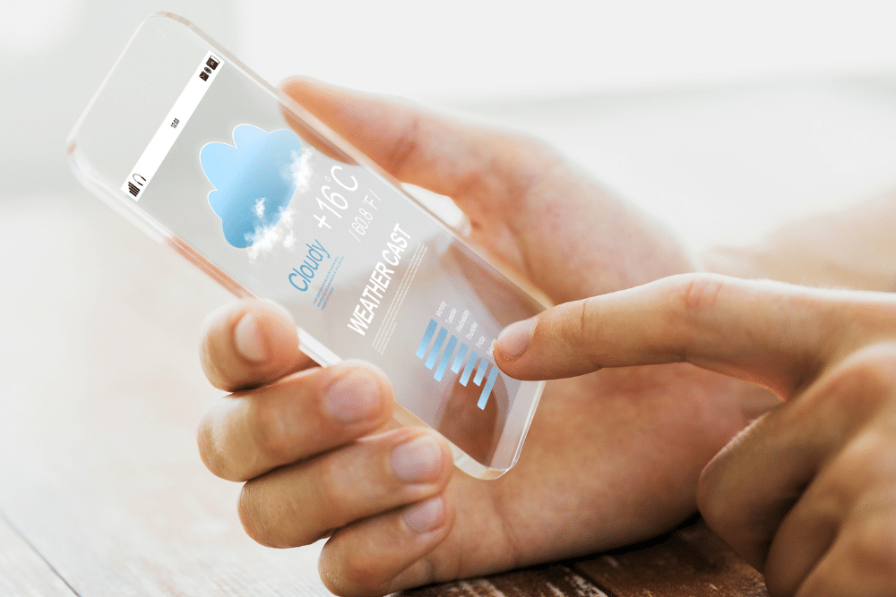 A pair of hands holds a transparent smartphone, upon which is displayed a generic weather app.